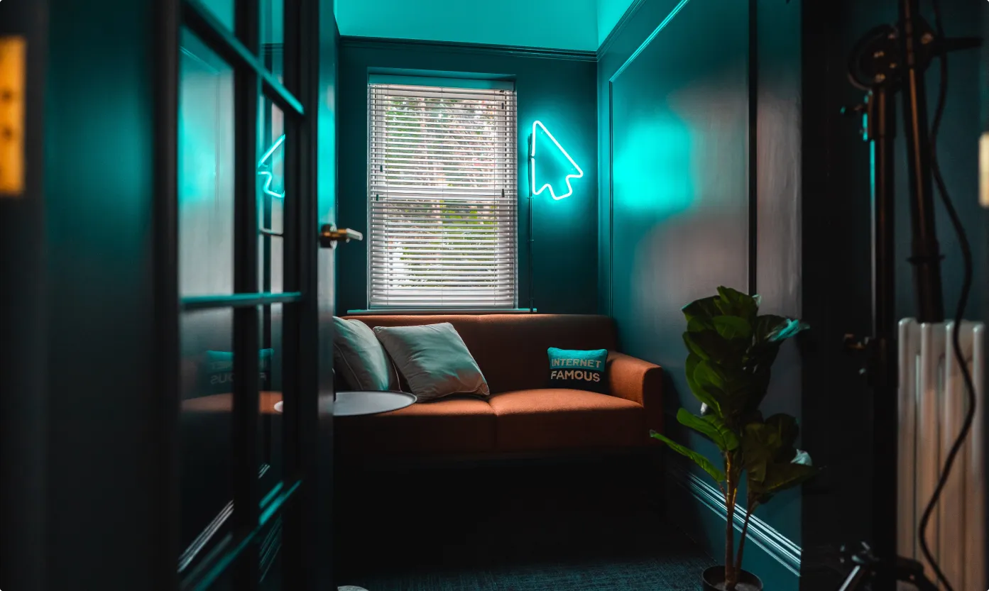 Cozy teal-lit room with brown sofa, pillows including one reading 'INTERNET FAMOUS', window blinds, and a potted plant.